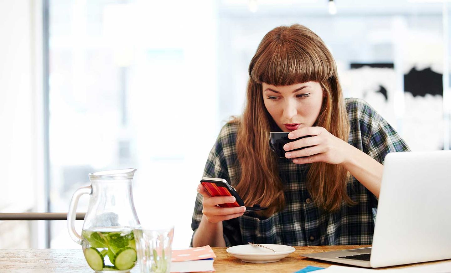 Eine Frau schaut auf ihr Smartphone und trinkt an einer Tasse