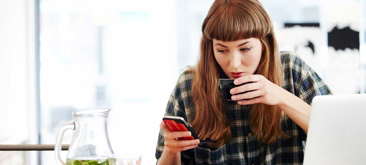 Eine Frau schaut auf ihr Smartphone und trinkt an einer Tasse