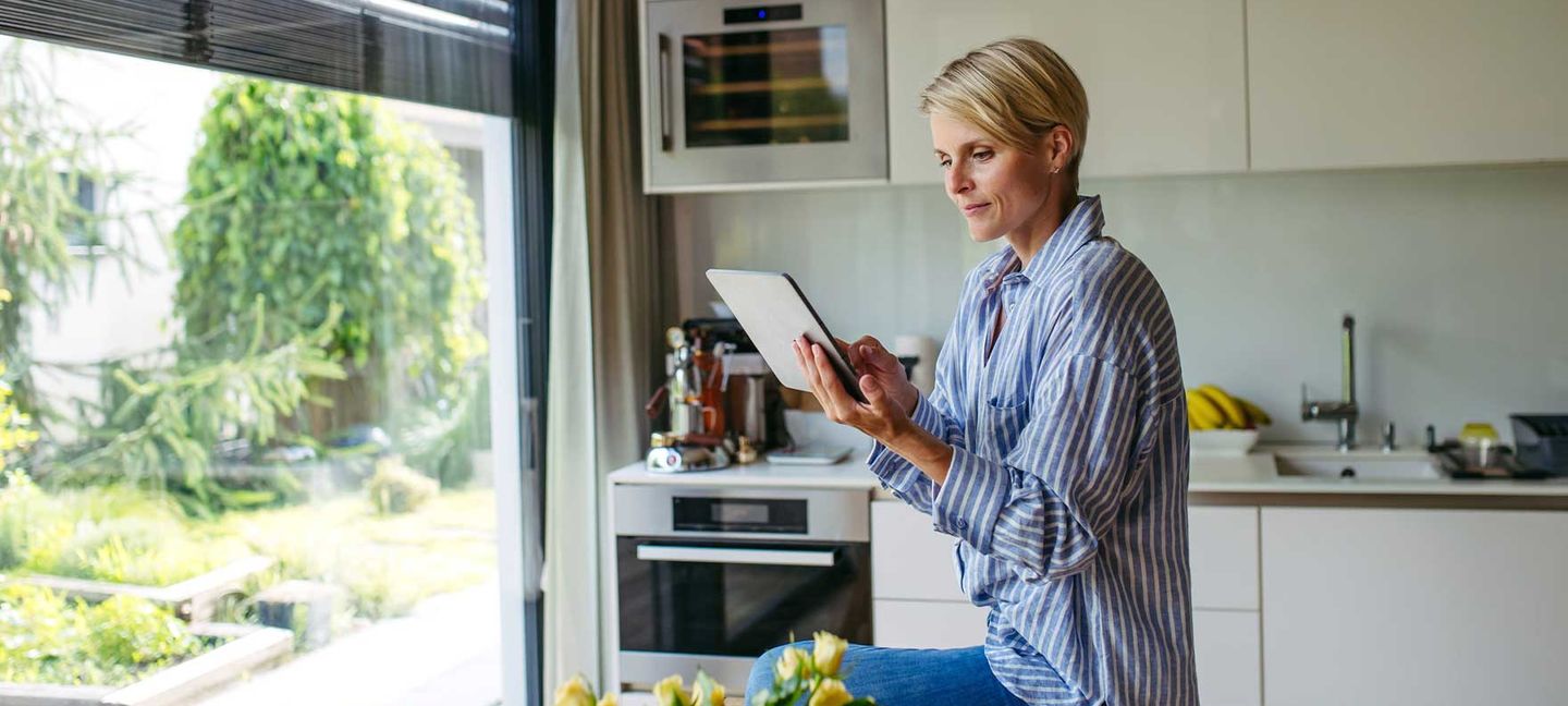 Eine junge Frau sitzt auf dem Küchentisch und informiert sich auf ihrem Tablet über die Verbrauchsrecherche. © Halfpoint Images / Moment via Getty Images