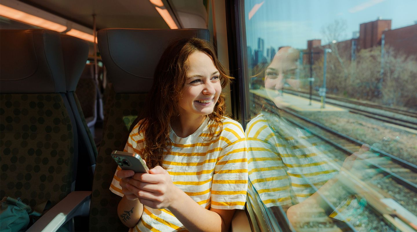 Eine junge Dame sitzt lächelnd im Zug mit einem Smartphone © Oleh_Slobodeniuk / E+ via Getty Images