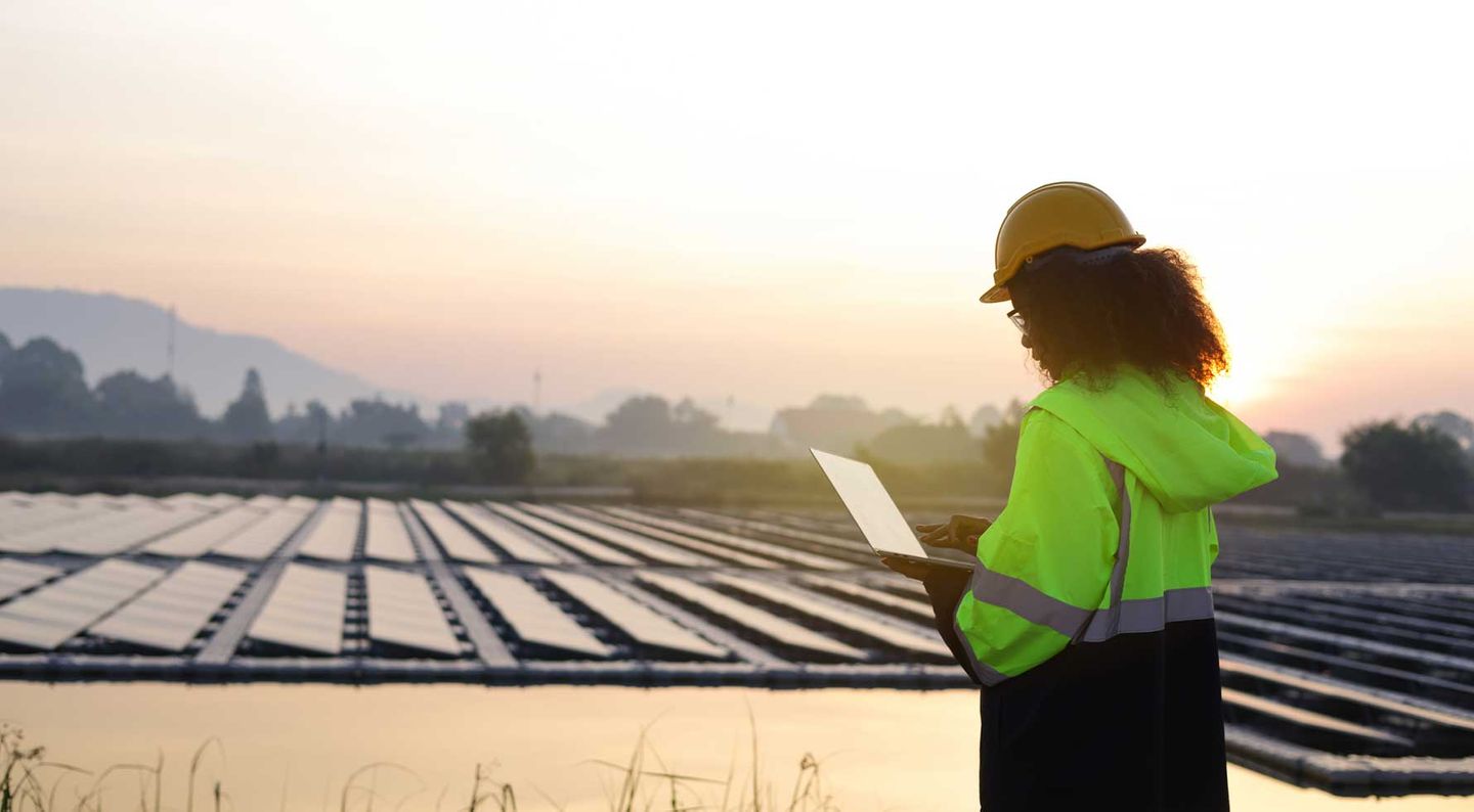 Eine Arbeiterin steht am Ufer eines schwimmenden Solarparks und überprüft die PV-Anlage am Laptop.
