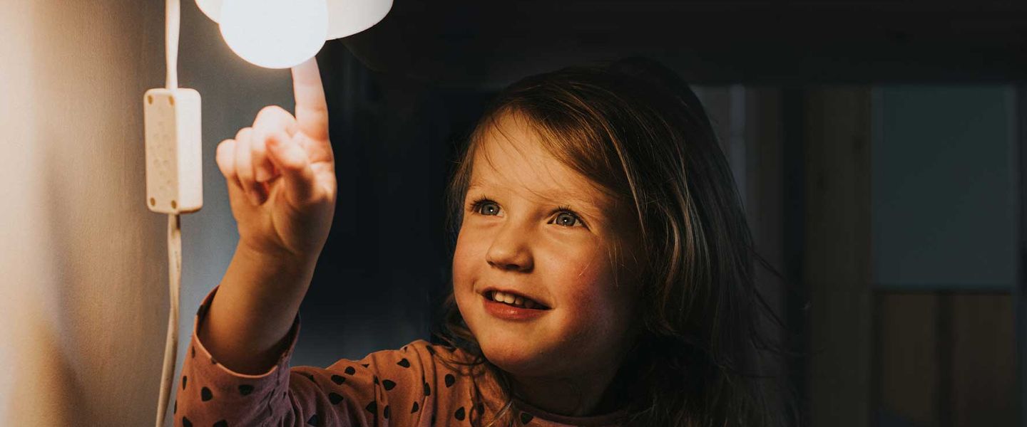 Ein kleines Mädchen sitzt vor einer eingeschalteten Lampe und berührt die Glühbirne mit dem Finger. © Catherine Falls Commercial / Moment via Getty Images