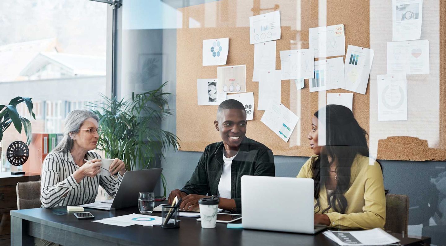 Zwei Frauen und ein Mann unterschiedlicher Generationen sitzen an einem Tisch an ihren Laptops und unterhalten sich. © PeopleImages / iStock / Getty Images Plus via Getty Images