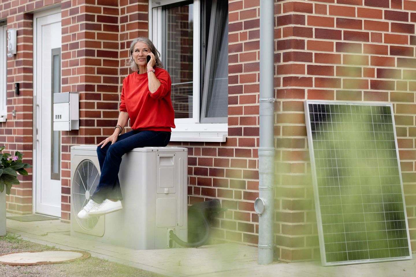 Eine Frau sitzt telefonierend auf einer Wärmepumpe vor Ihrem Haus. © Westend61 / Westend61 via Getty Images