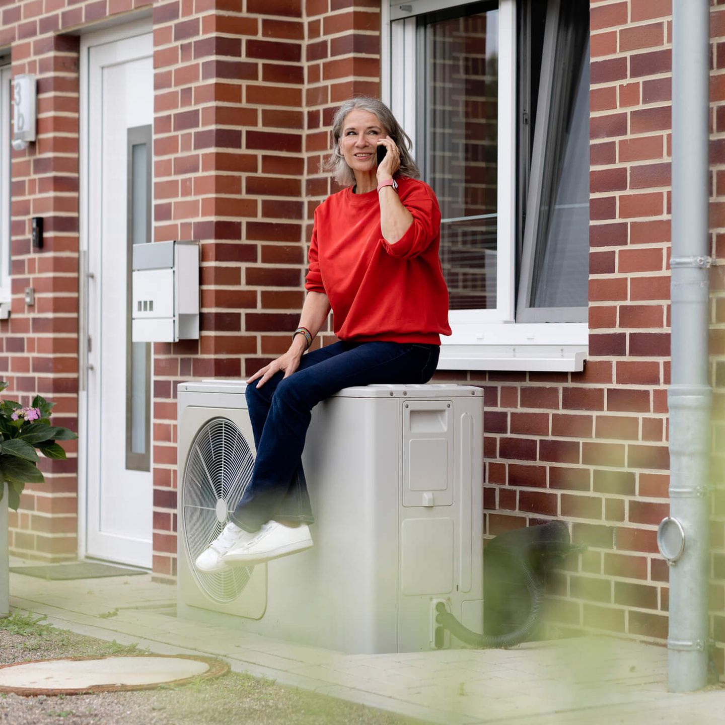 Frau sitzt telefonierend auf einer Wärmepumpe vor Ihrem Haus. © Westend61 / Westend61 via Getty Images