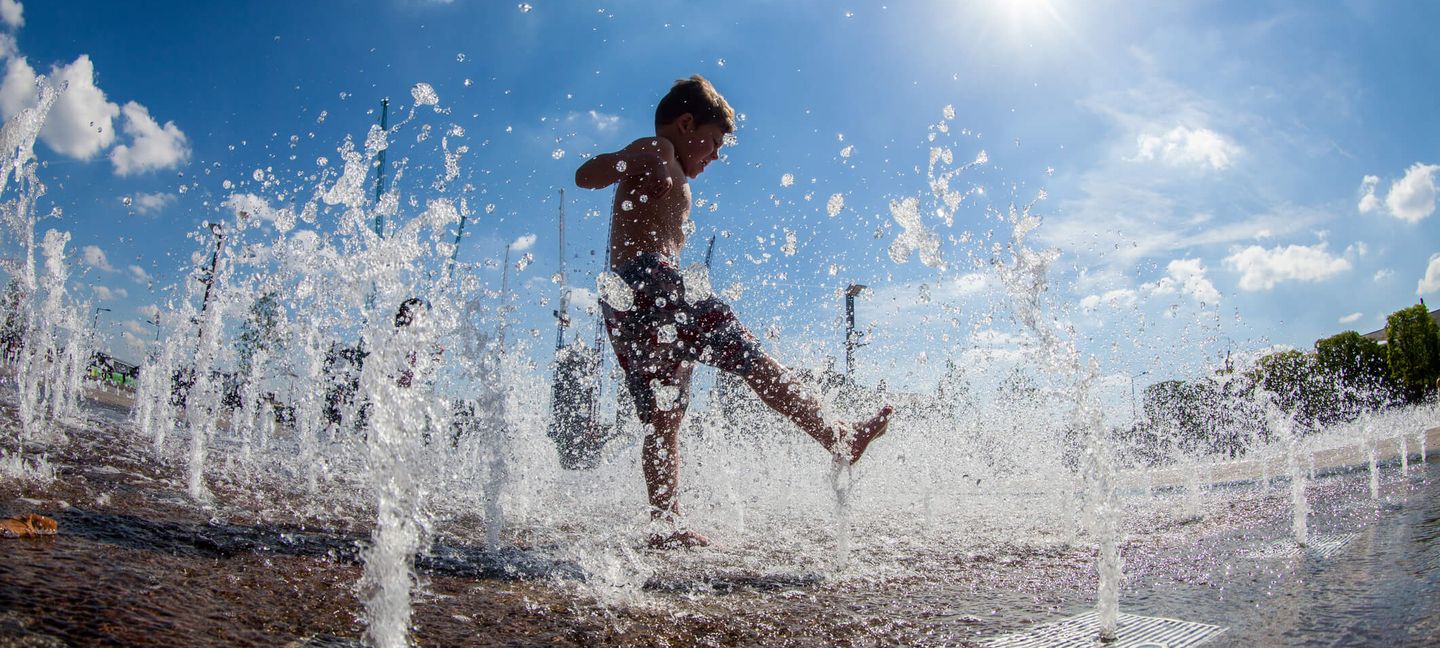 Ein Kind spielt in sprudelnden Wasserfontänen auf einem Platz bei strahlendem Sommerwetter. © paul mansfield photography / Moment via Getty Images © paul mansfield photography / Moment via Getty Images