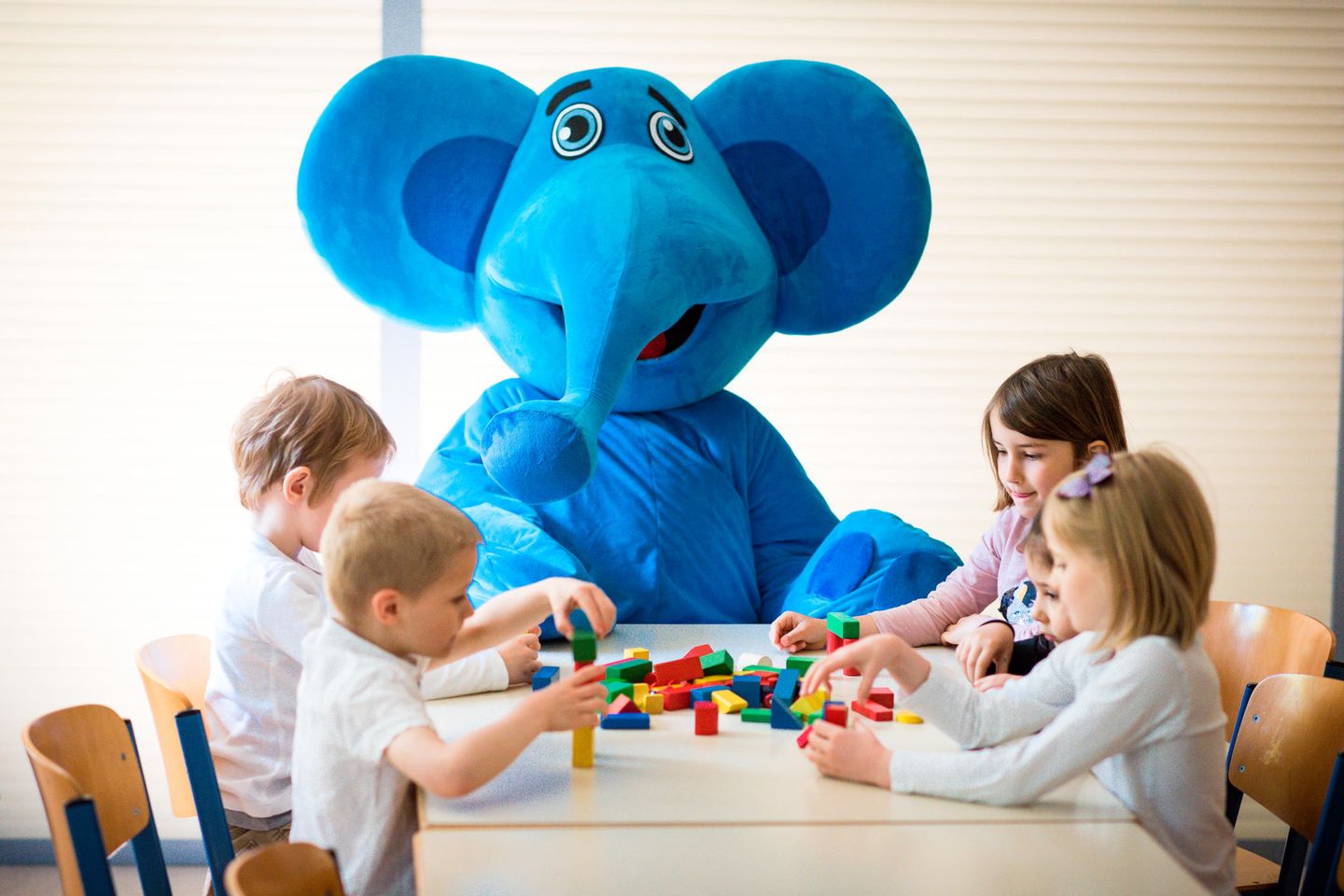 Das Maskottchen des KSB im blauen Elefantenkostüm sitzt mit spielenden Kindern an einem Tisch.