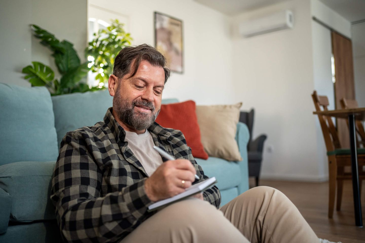 Ein Mann sitzt vor seinem Sofa auf dem Boden und schreibt in ein Notizbuch, im Hintergrund hängt ein Klimagerät an der Wand. © Milan Markovic via Getty Images