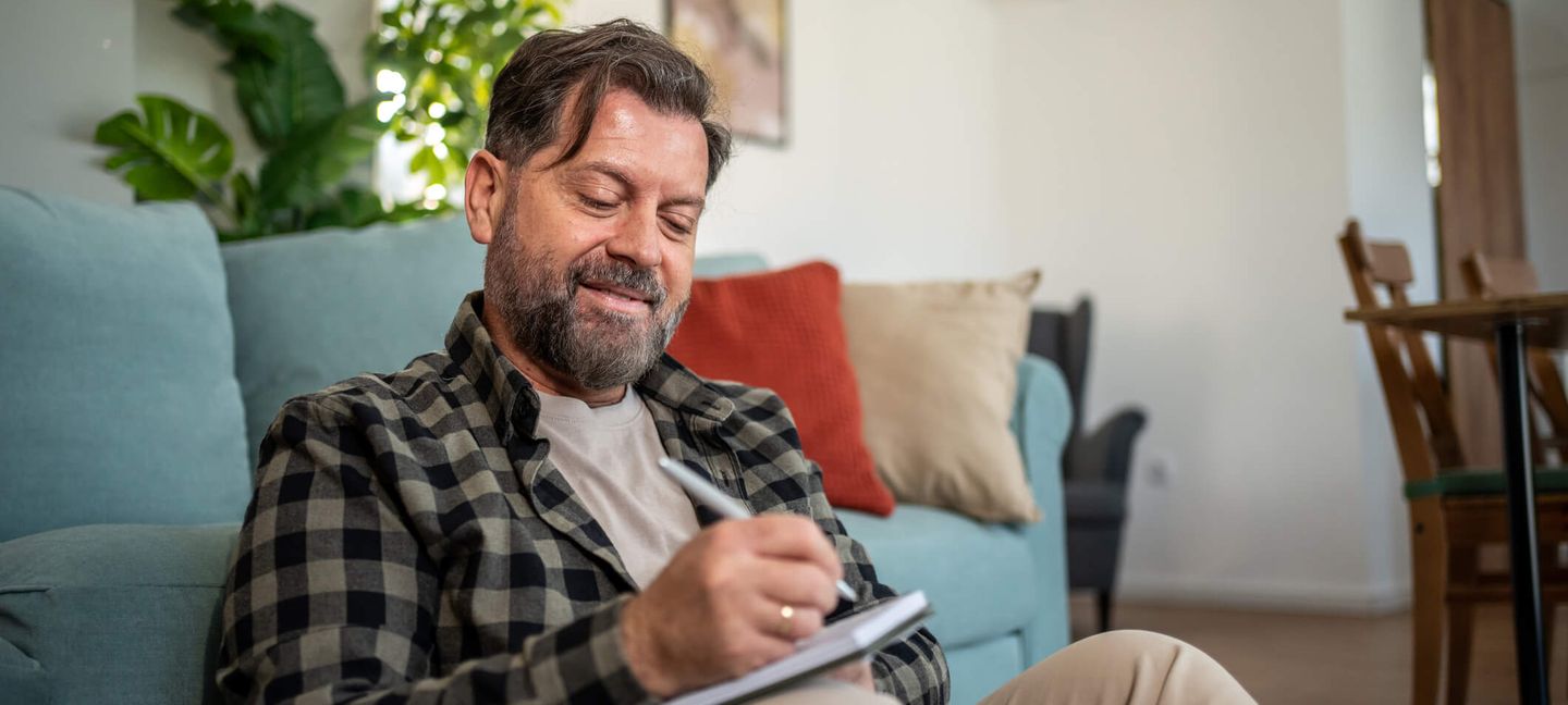 Ein Mann sitzt vor seinem Sofa auf dem Boden und schreibt in ein Notizbuch, im Hintergrund hängt ein Klimagerät an der Wand. © Milan Markovic via Getty Images