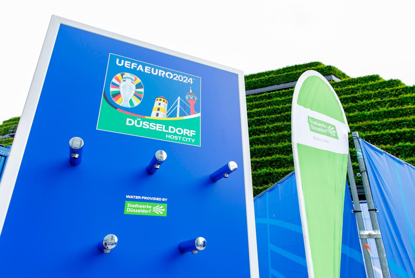 Ein Trinkwasserspender mit Logo der UEFA EURO 2024 steht vor einer begrünten Fassade in Düsseldorf. 