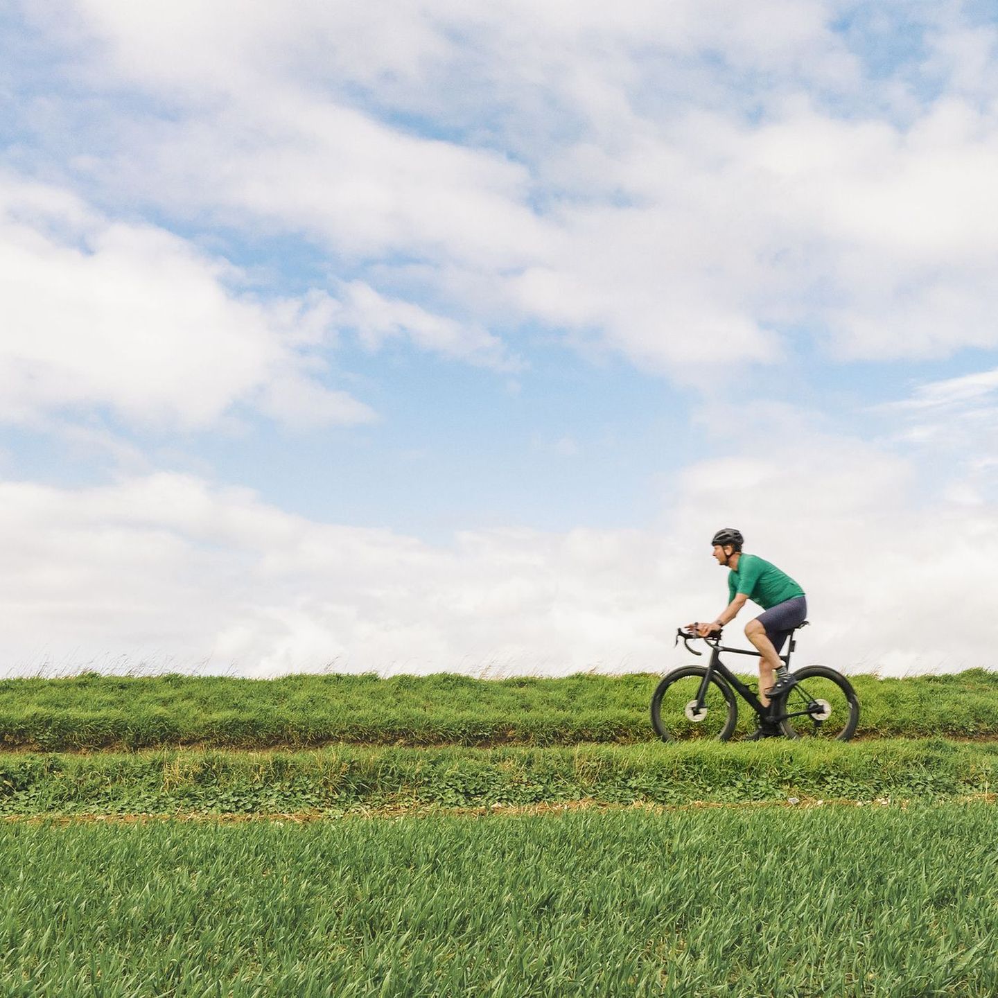 Ein Radfahrer in grüner Kleidung fährt auf einem Feldweg entlang, umgeben von grünen Feldern und blauem Himmel. © Justin Paget / DigitalVision via Getty Images