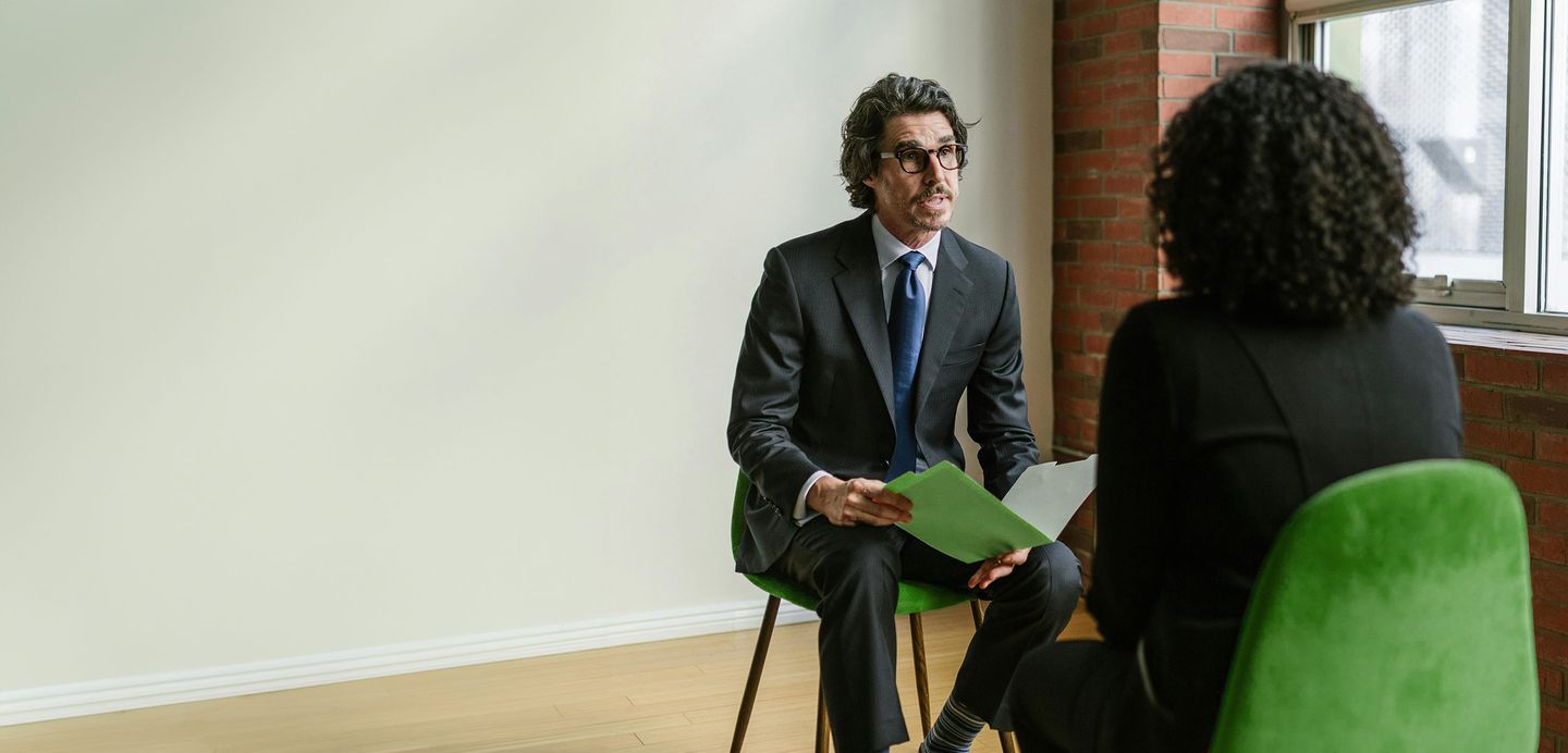Ein Berater im Anzug sitzt auf deinem grünen Stuhl und unterhält sich mit einer Frau Foto von RDNE Stock project via https://www.pexels.com/photo/a-man-in-black-suit-sitting-on-a-yellow-chair-8124241/