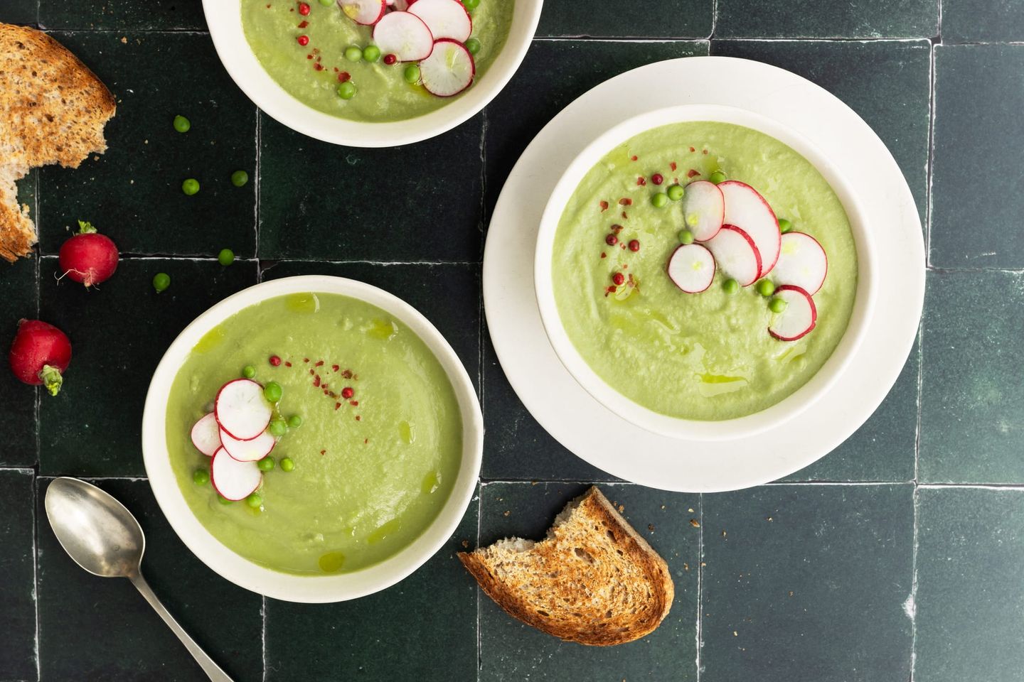 Drei Schüsseln mit grüner Erbsensuppe, garniert mit Radieschenscheiben und rosa Pfeffer, serviert mit geröstetem Brot