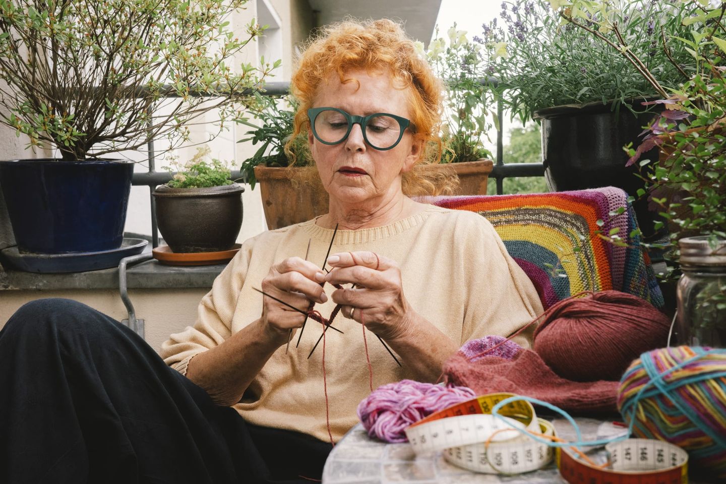 Ältere Frau mit roten Locken und grüner Brille strickt konzentriert auf einem Balkon. Umgeben von Blumentöpfen, Wollknäueln, einem Maßband und einem bunten Häkelkissen im Hintergrund.