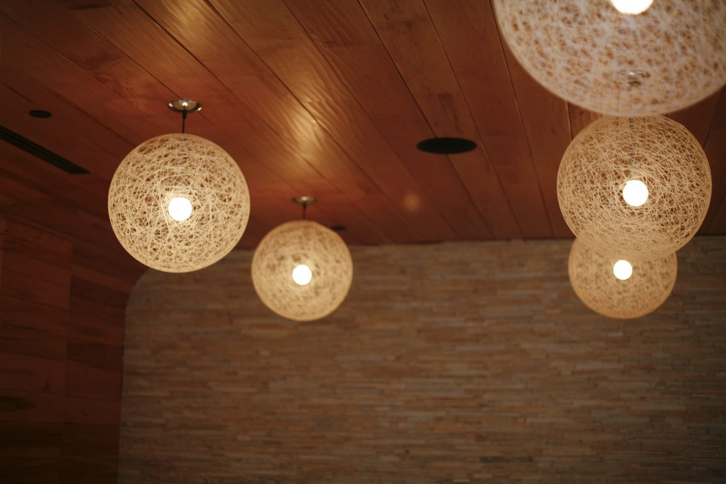 Decke mit mehreren runden Pendelleuchten aus filigranem Fadengeflecht, die warmes Licht in einem Raum mit Holzdecke und gemauerter Wand spenden