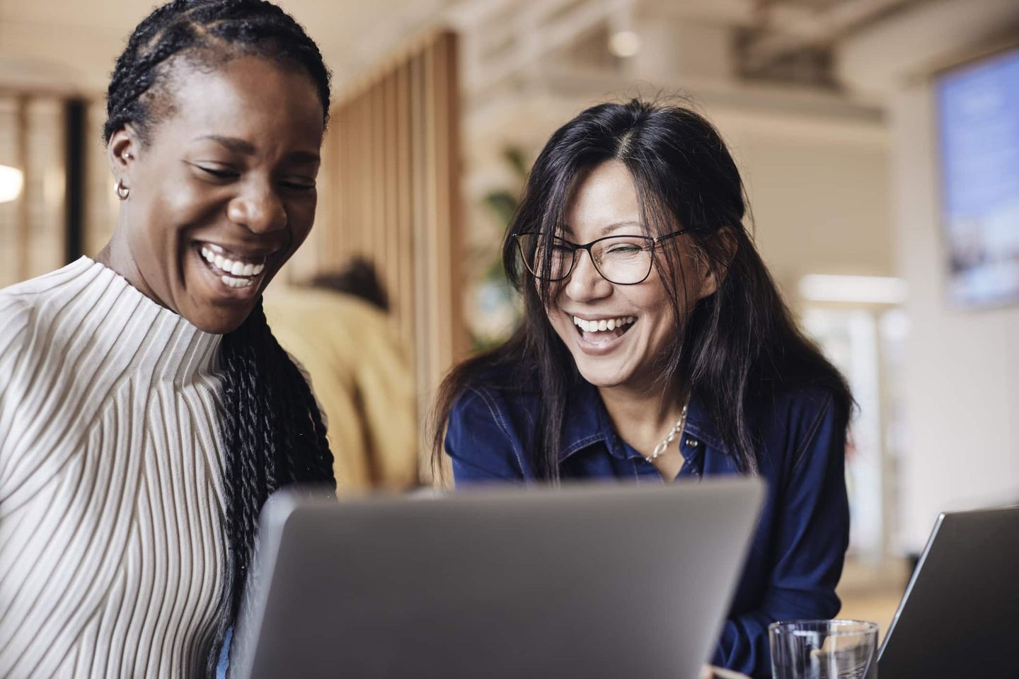 Zwei Frauen lachen gemeinsam vor einem Laptop in einem offenen Arbeitsbereich. Getty