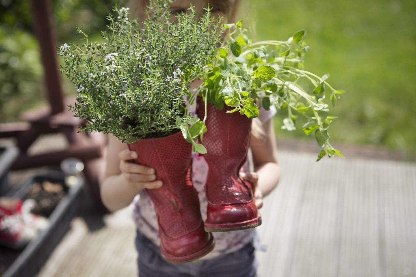 Ein Kind hält zwei rote Gummistiefel, die als kreative Pflanzgefäße für Kräuter wie Thymian und Oregano genutzt werden.
