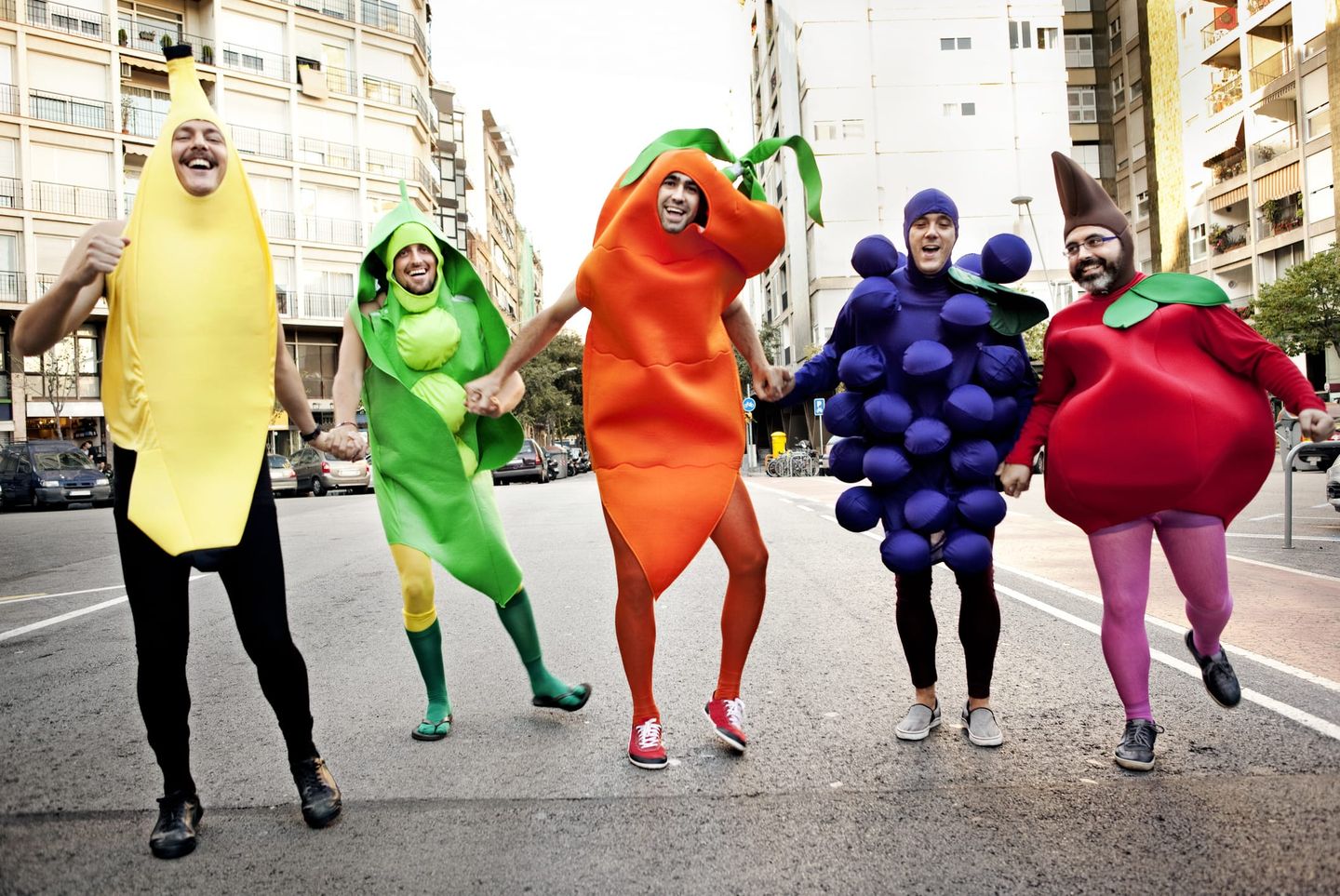 Eine fröhliche Gruppe Erwachsener, verkleidet als Banane, Erbse, Karotte, Traube und Kirsche, tanzt während eines Karnevalsumzugs auf der Straße.