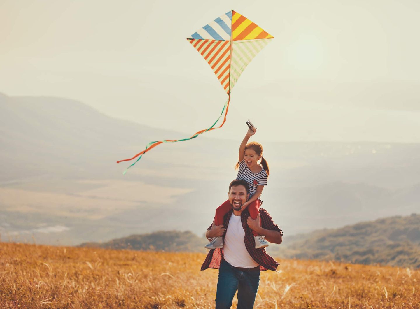 Ein Vater trägt seine Tochter auf den Schultern, während sie gemeinsam einen gestreiften Drachen steigen lassen. Getty