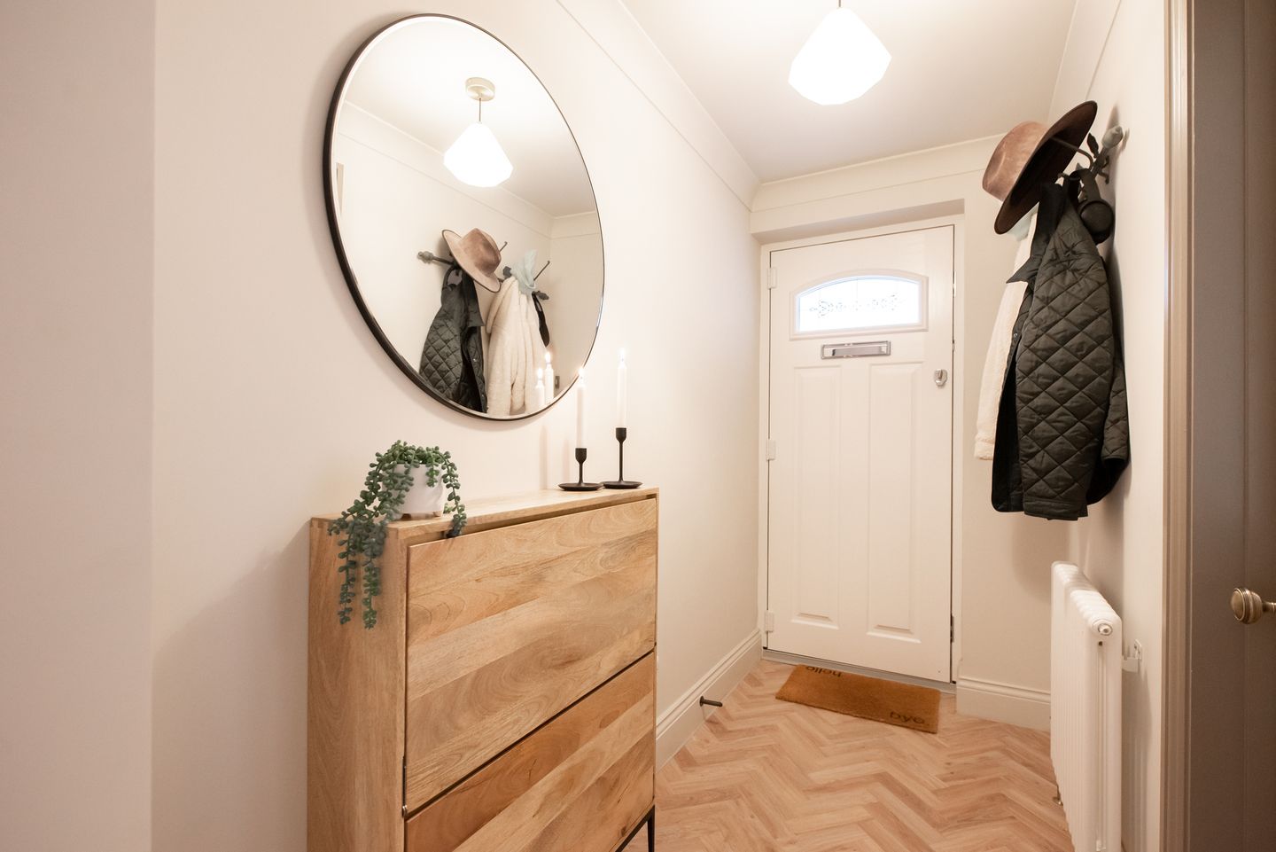 Heller Eingangsbereich mit einer Holzkommode, einem runden Wandspiegel, zwei Kerzenhaltern und einer kleinen Zimmerpflanze; an der Wand gegenüber hängt eine Garderobe mit Jacken und einem Hut.