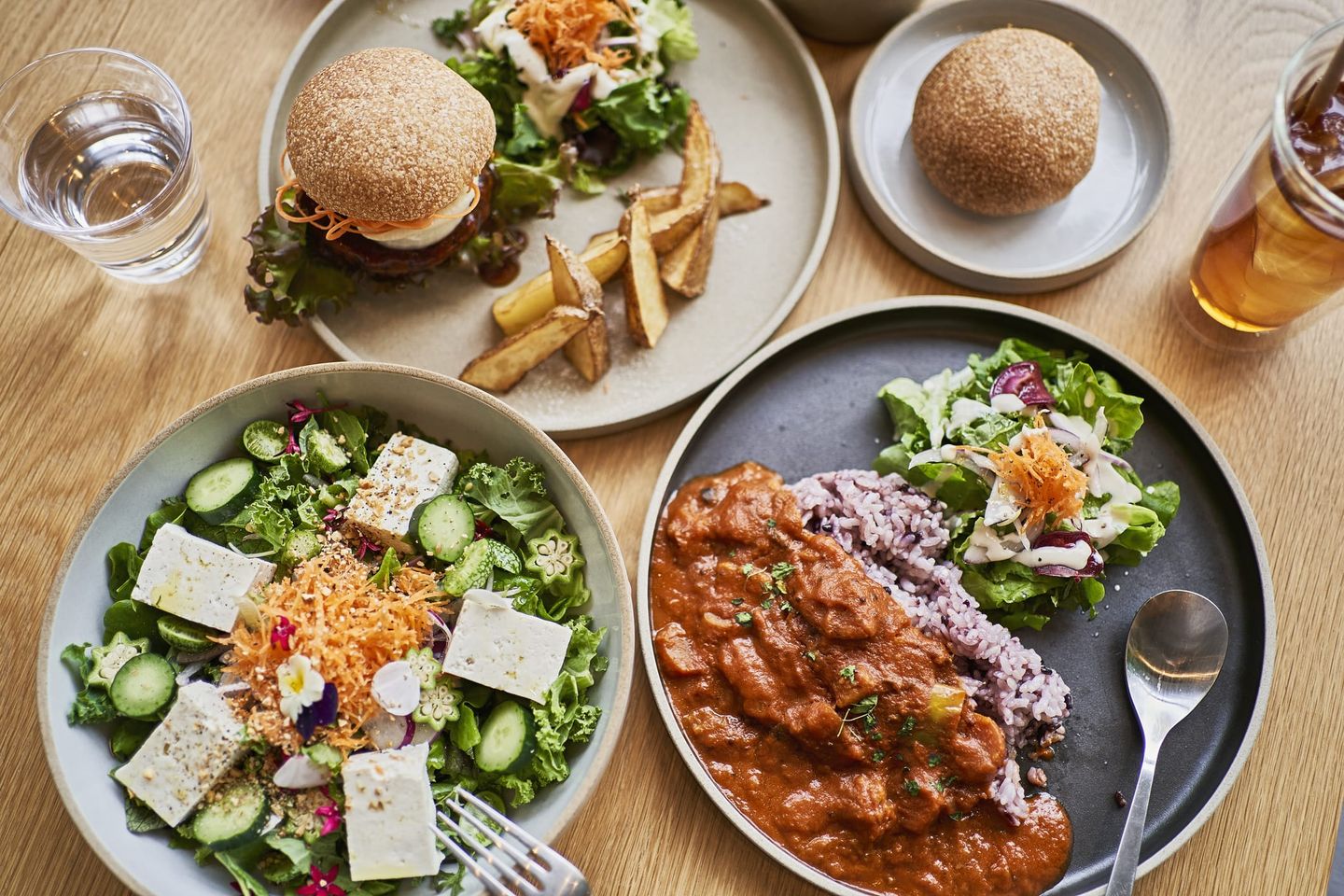 Vielfältige vegane Mahlzeiten auf einem Holztisch, darunter ein Burger mit Pommes, Salat mit Tofu, ein Teller mit Curry und Reis sowie Wasser und Eistee