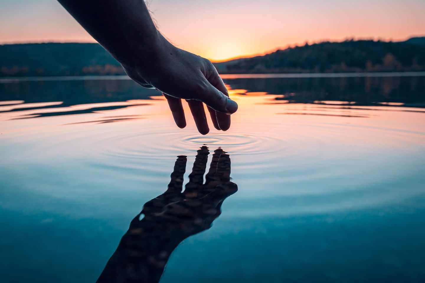 Hand berührt Wasseroberfläche bei Sonnenuntergang, Spiegelung im Wasser sichtbar Getty