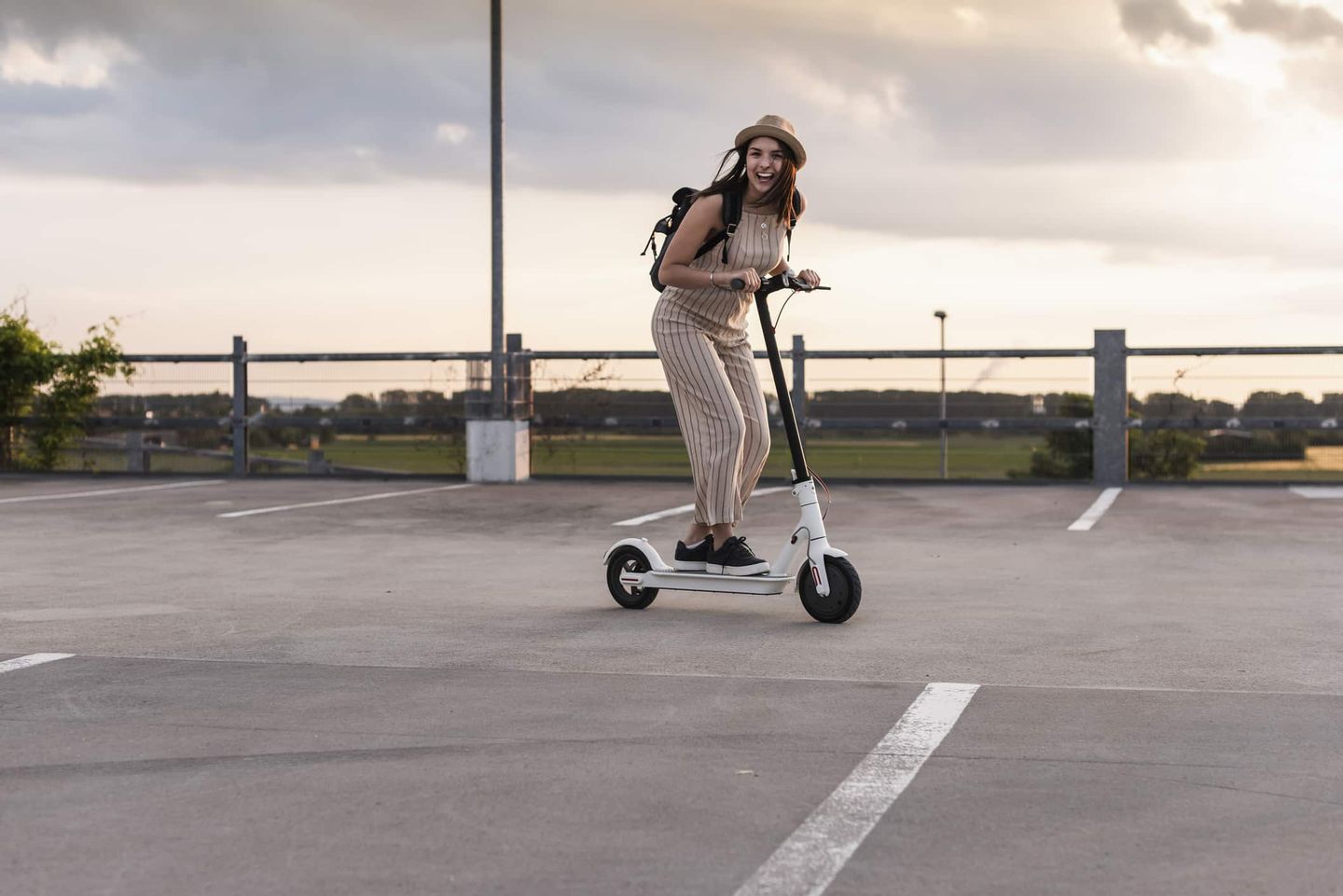 Eine junge Frau fährt lachend mit einem E-Scooter auf einem offenen Parkplatz bei Sonnenuntergang. Getty