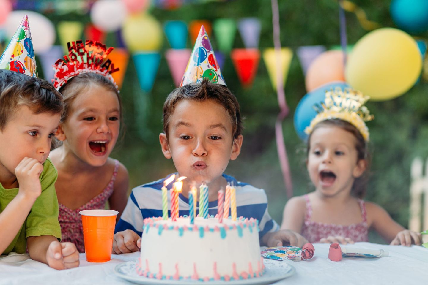 Ein Junge mit Partyhut bläst die Kerzen auf einer Geburtstagstorte aus, umgeben von fröhlich jubelnden Kindern.