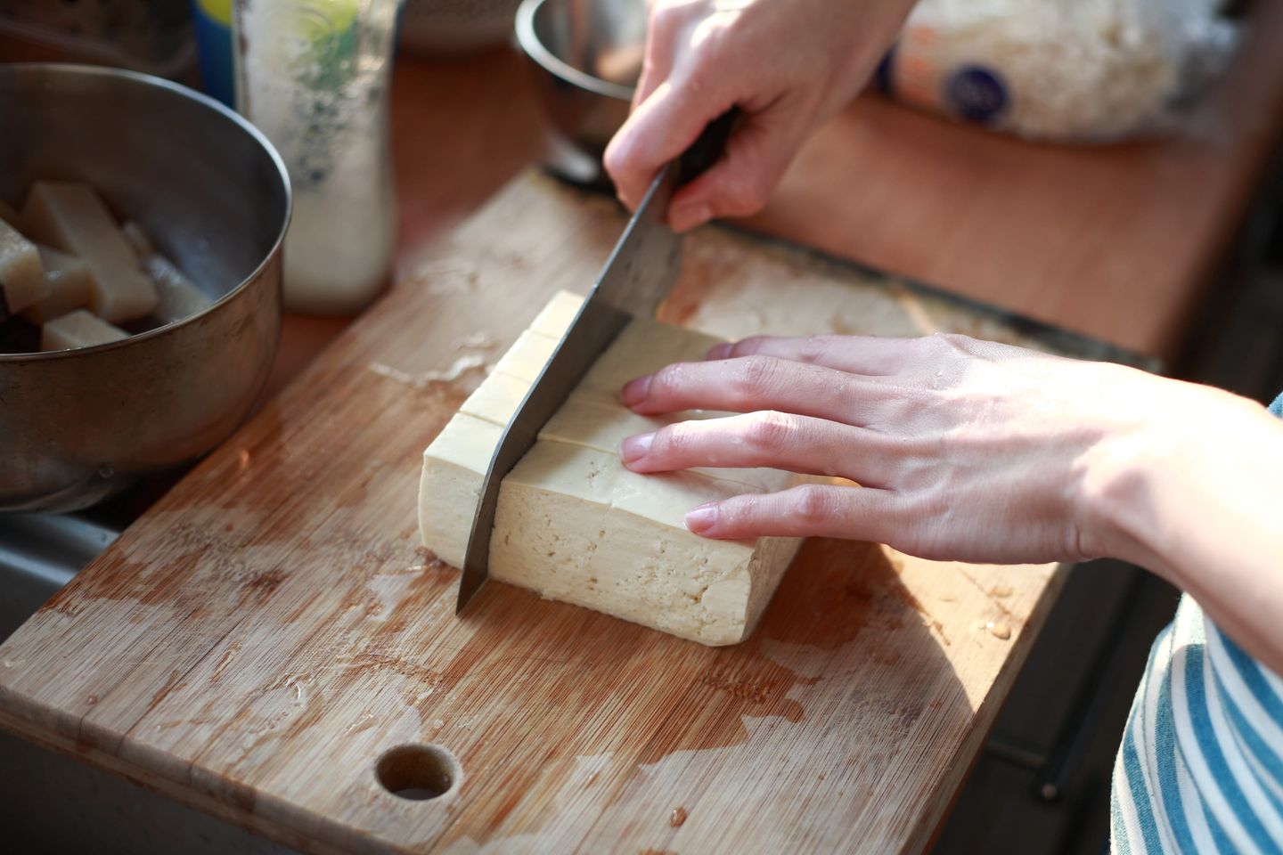 Eine Hand schneidet frischen Tofu auf einem Holzbrett in Würfel, mit Küchengeräten und Zutaten im Hintergrund