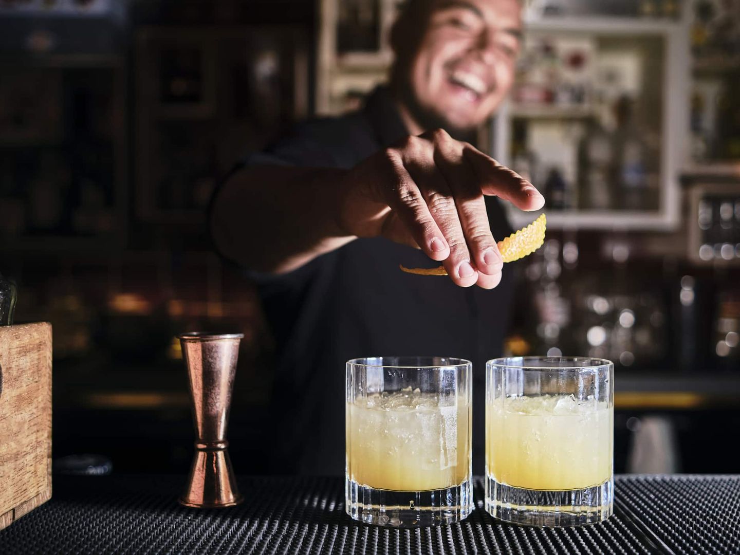 Ein Barkeeper garniert zwei gelbe Cocktails mit einer Zitronenzeste und lacht dabei. Getty