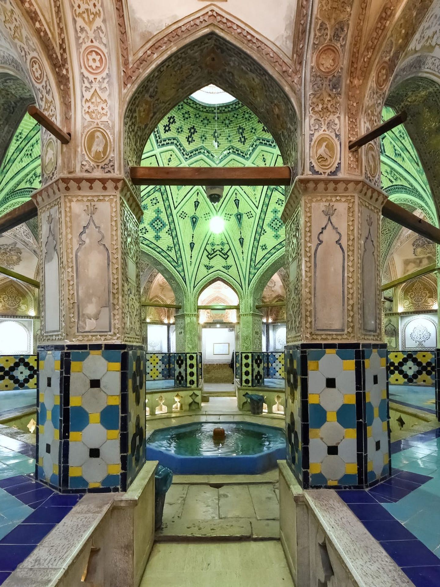 Innenansicht eines historischen Hammams mit reich verzierten, farbenfrohen Mosaiken, hohen Gewölbedecken und einem zentralen Brunnen unter grünen Kuppeln