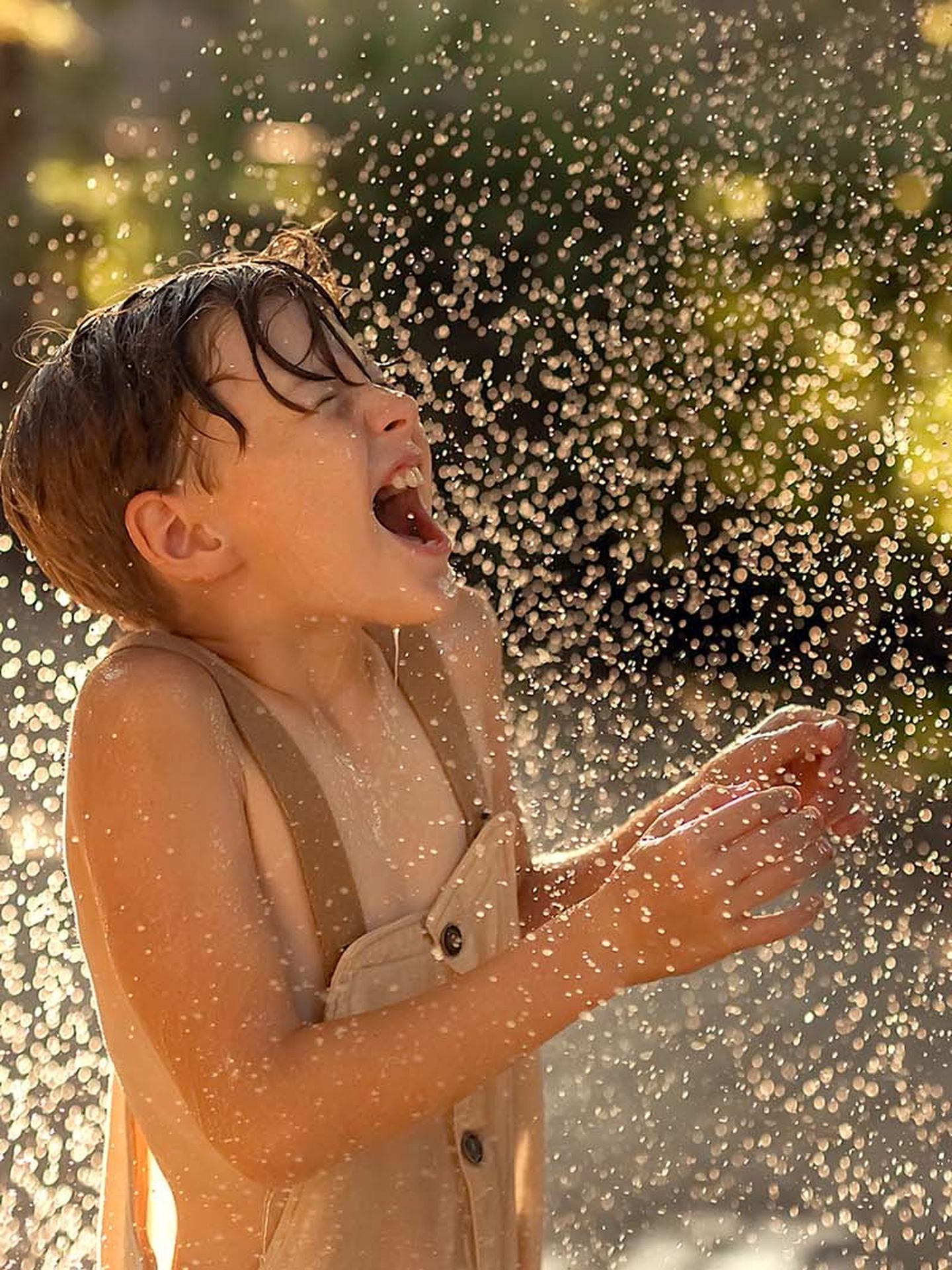 Ein lachendes Kind spielt im Freien mit Wasser. Das Wasser spritzt in feinen Tropfen in die Luft, während das Kind, durchnässt und mit geschlossenen Augen, den Moment sichtlich genießt.