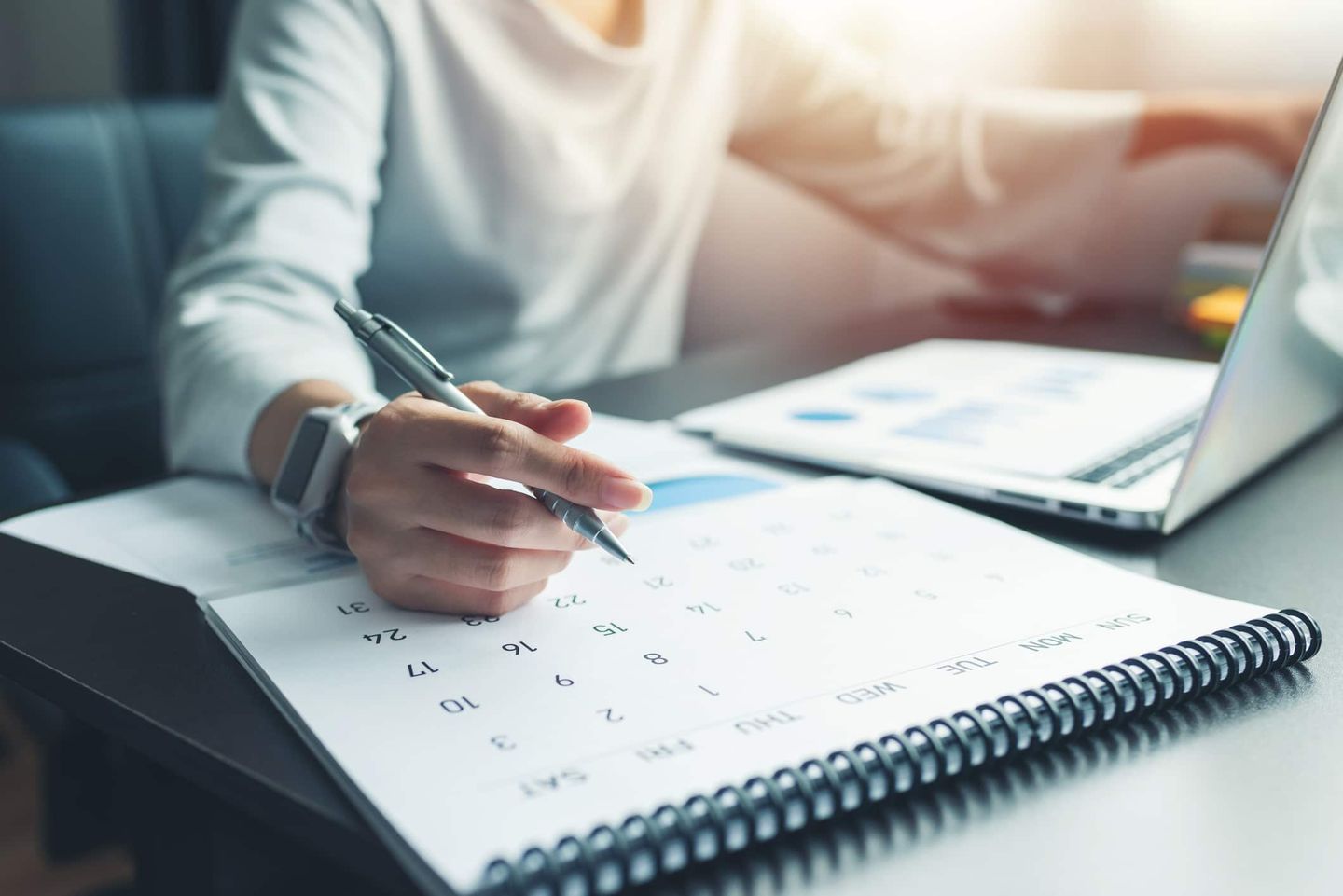 Hand mit Stift über großem Kalender neben Laptop. Getty