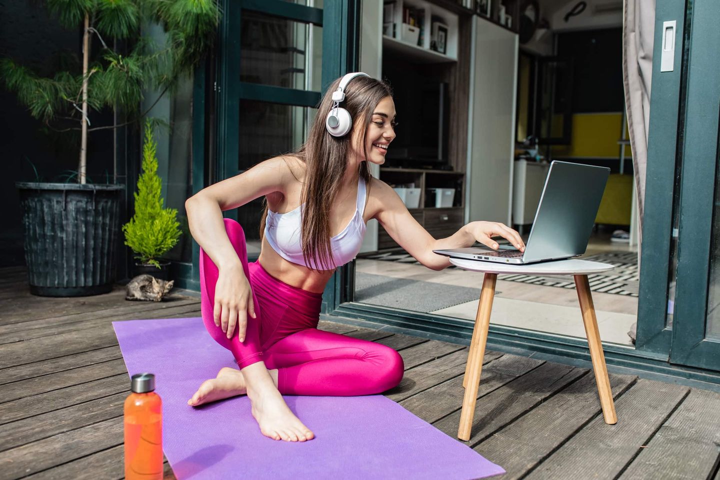 Junge Frau in Sportkleidung mit Kopfhörern macht Online-Yoga auf der Terrasse Getty