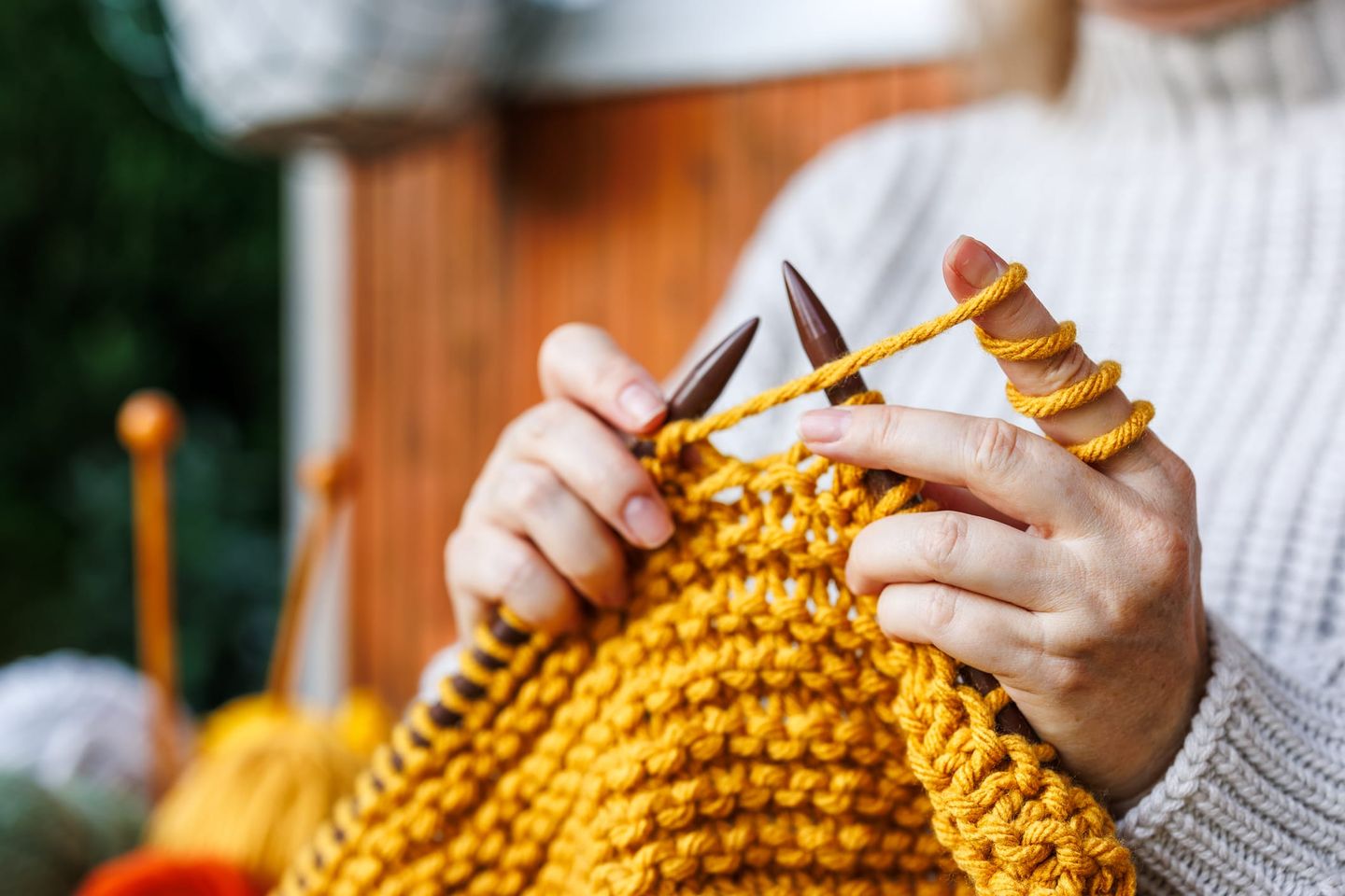 Nahaufnahme zweier Hände, die mit dicken Stricknadeln aus Holz ein grobes Strickstück aus senfgelber Wolle stricken. Die Person trägt einen cremefarbenen Strickpullover.
