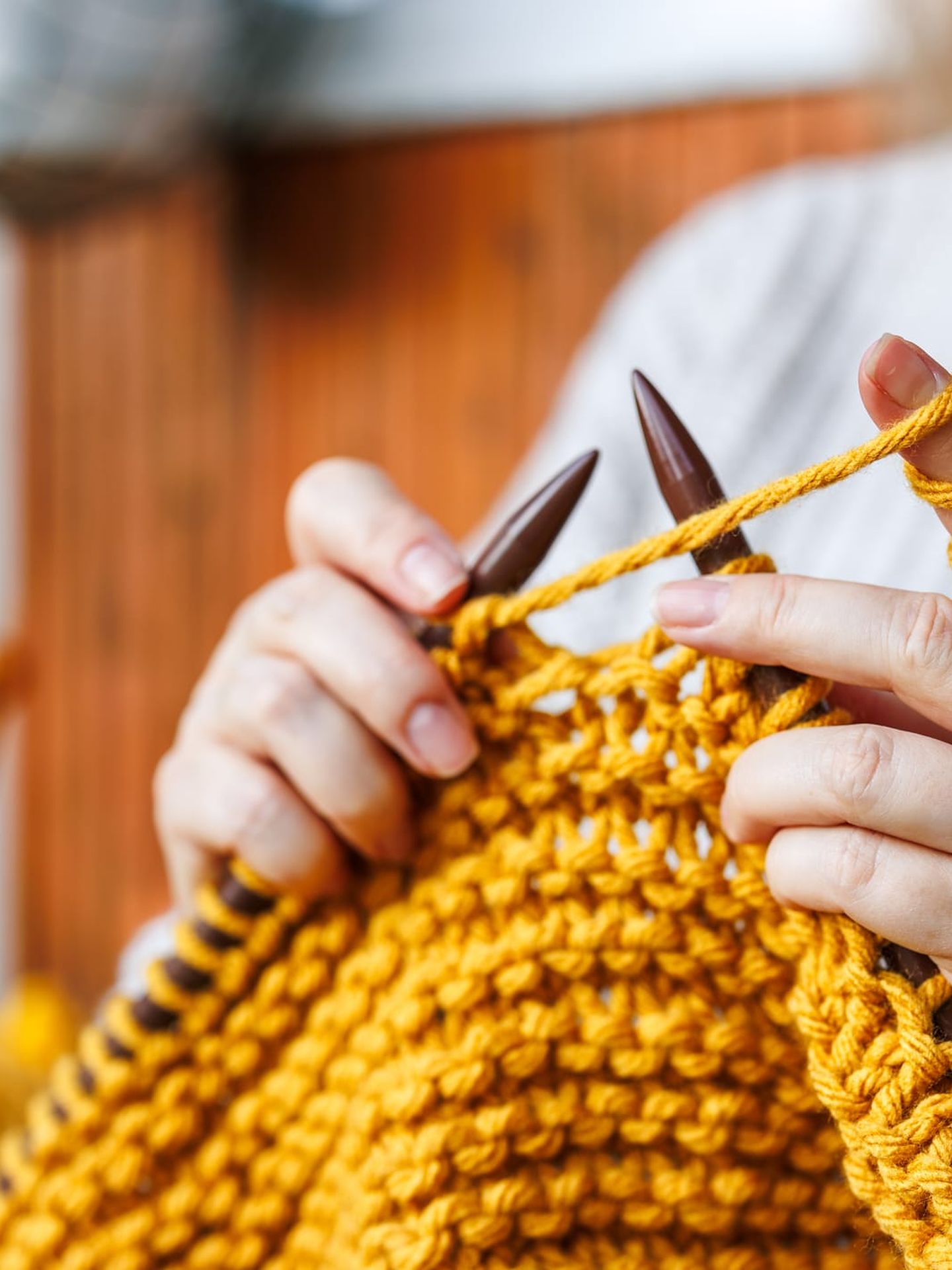 Nahaufnahme zweier Hände, die mit dicken Stricknadeln aus Holz ein grobes Strickstück aus senfgelber Wolle stricken. Die Person trägt einen cremefarbenen Strickpullover.