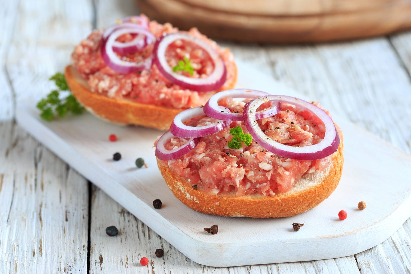 Ein Brötchen mit rohem Mett, garniert mit roten Zwiebelringen und Petersilie, auf einem weißen Holzbrett.