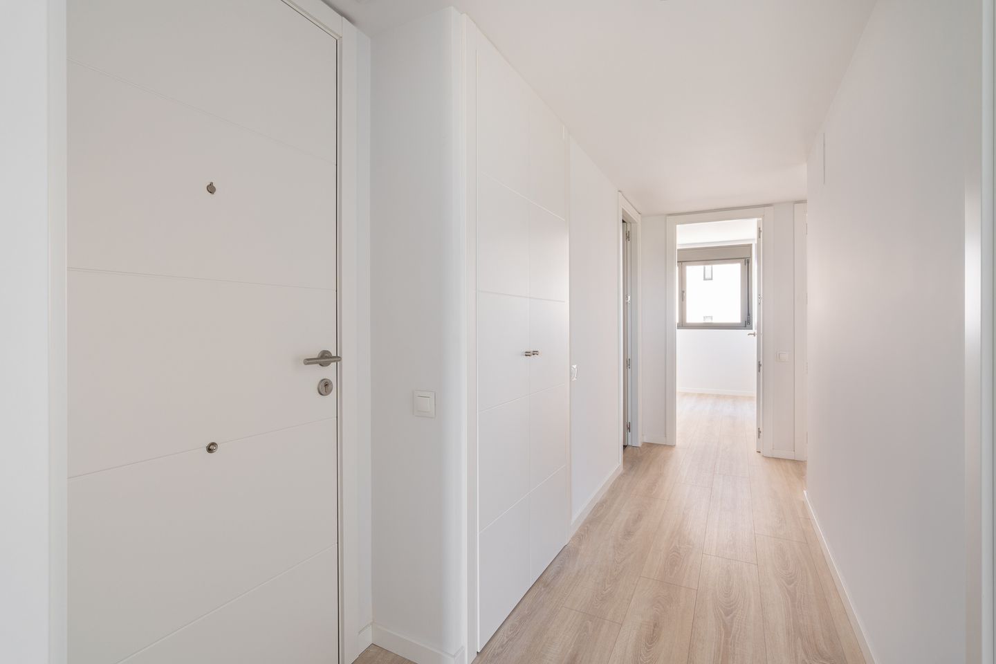 Heller, minimalistischer Flur mit weißen Türen, Wänden und Holzfußboden, der zu einem Raum mit großem Fenster führt