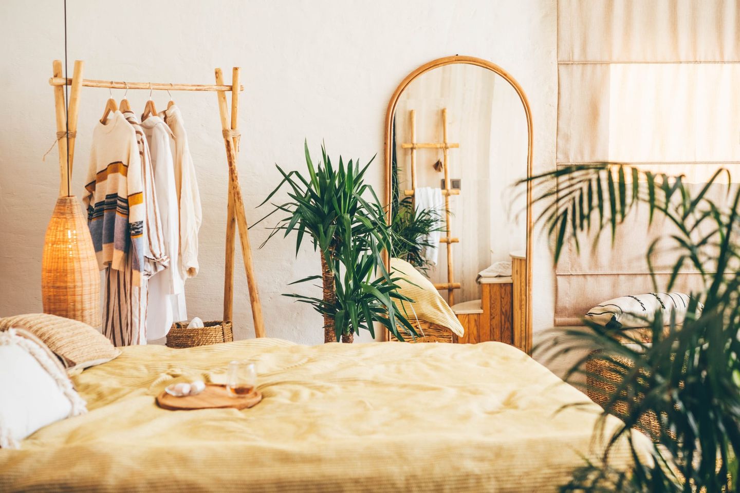 Gemütliches Schlafzimmer im Boho-Stil mit großem Spiegel, Zimmerpflanzen, gelber Bettdecke und Holzkleiderständer mit hängender Kleidung.