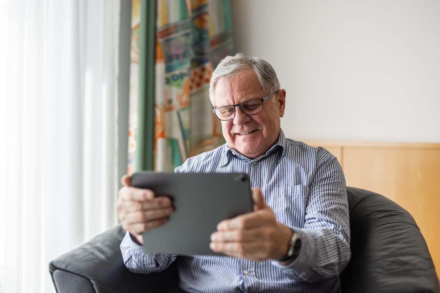 Ein älterer Mann sitzt auf einem Sessel und lächelt, während er ein Tablet benutzt. Getty