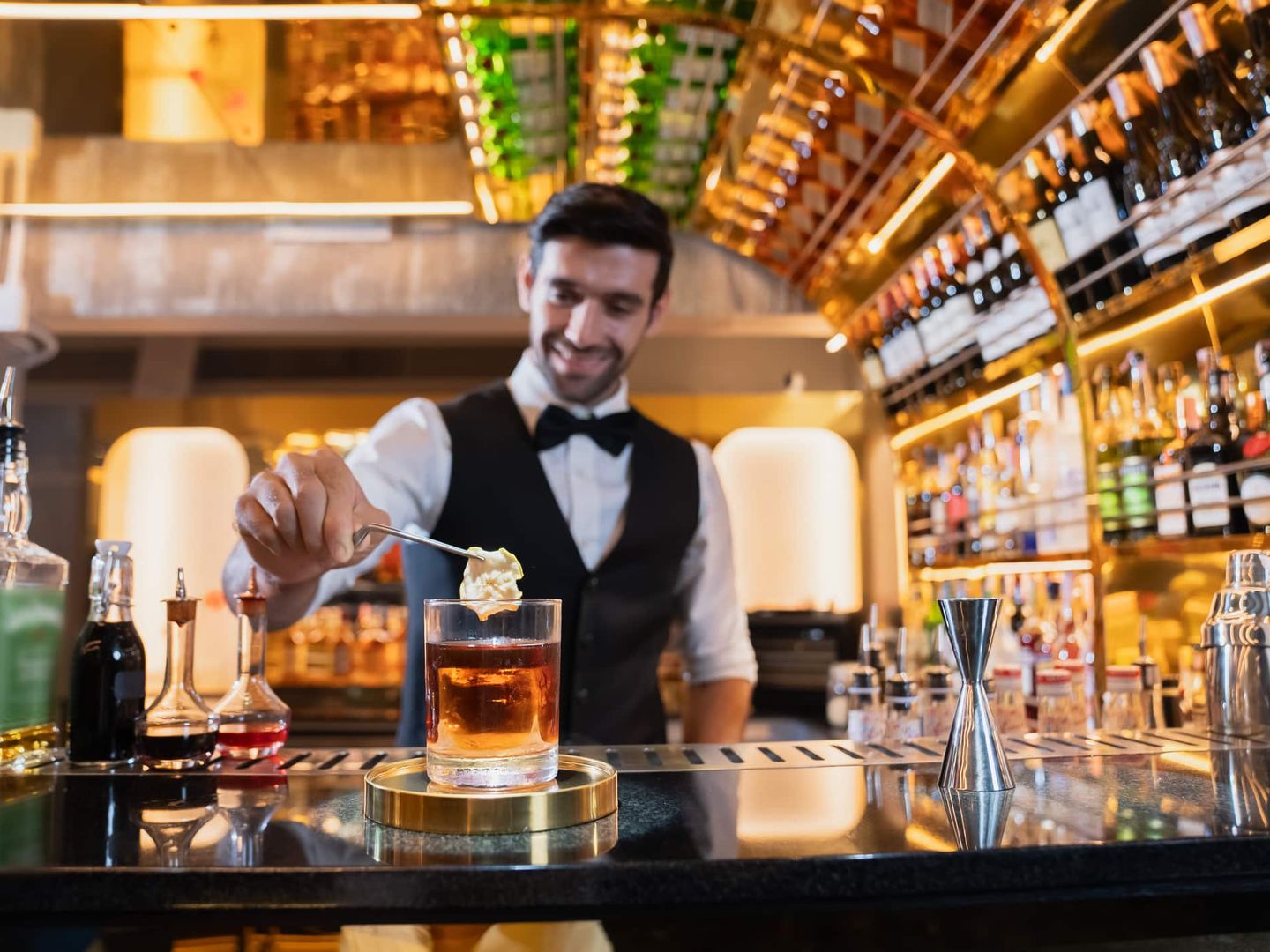 Ein Barkeeper im eleganten Anzug bereitet an einer beleuchteten Bar einen Cocktail mit Eis zu. Getty