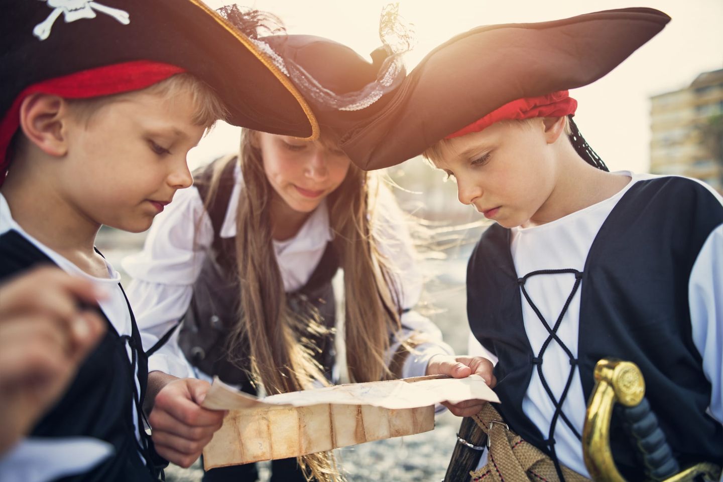 Drei Kinder in Piratenkostümen studieren gemeinsam eine Schatzkarte bei Sonnenschein.