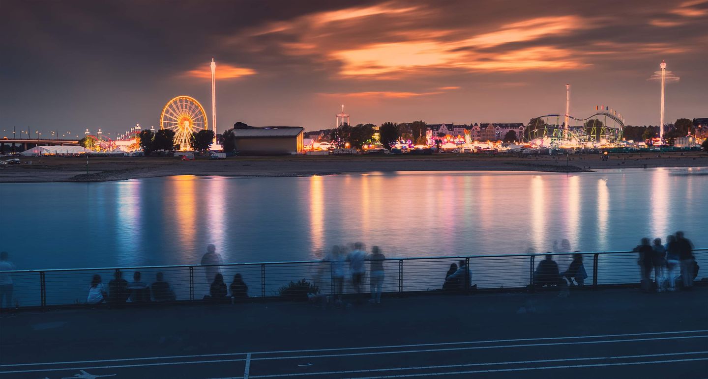 Blick auf die Rheinkirmes Düsseldorf mit beleuchtetem Riesenrad und Fahrgeschäften bei Sonnenuntergang am Rhein Getty
