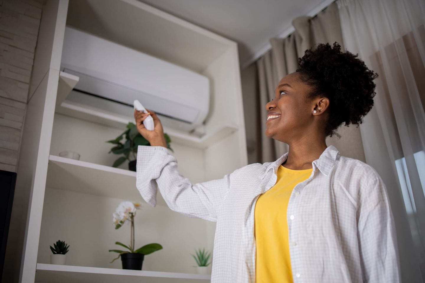 Frau steht im Wohnzimmer und bedient mit einer Fernbedienung die Wand-Klimaanlage, lächelt zufrieden.