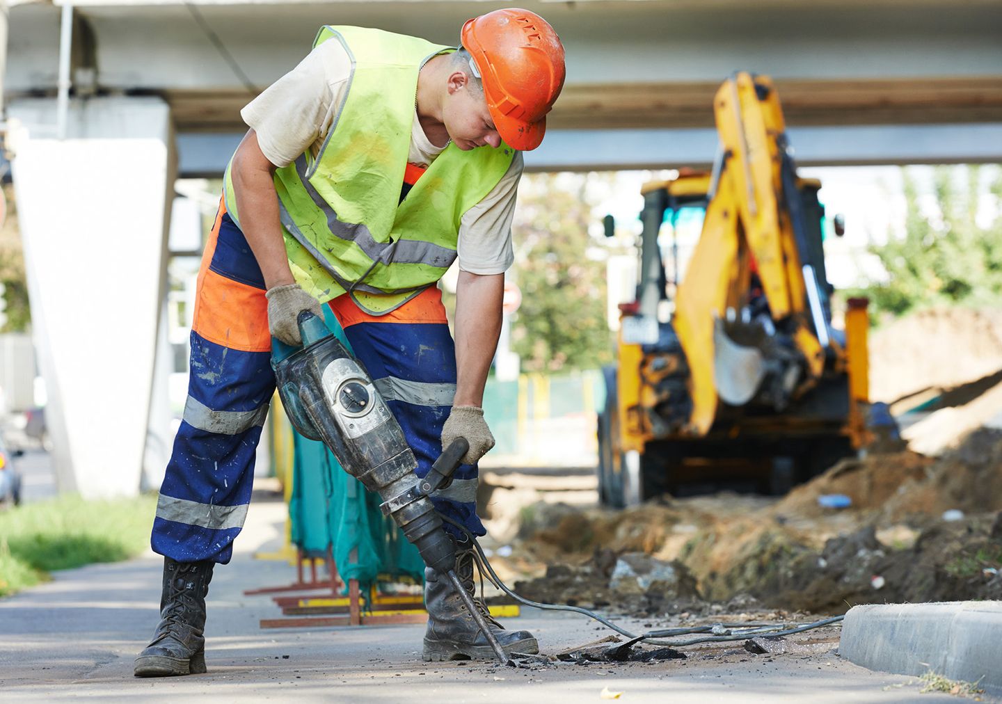 Ein Bauarbeiter in Warnweste und Helm arbeitet mit einem Presslufthammer auf einer Baustelle, im Hintergrund ist ein Bagger zu sehen.