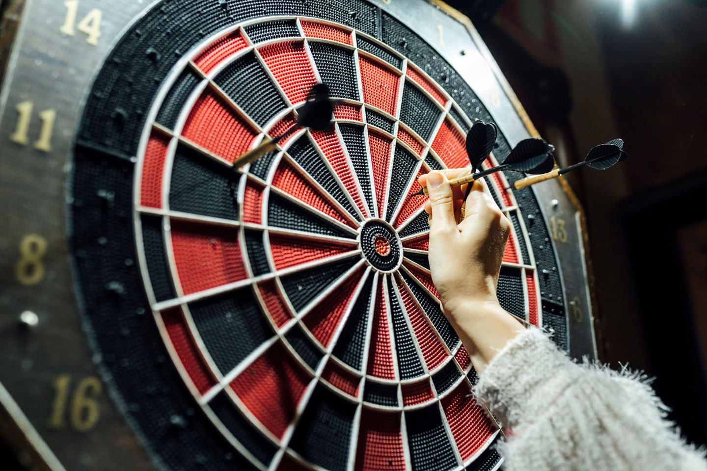 Eine Hand zieht einen schwarzen Dartpfeil aus einer elektronischen Dartscheibe, zwei weitere Pfeile stecken bereits darin.