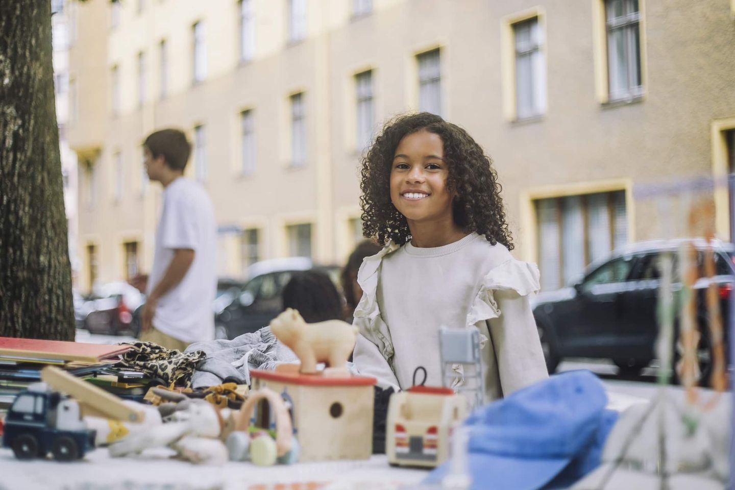 Mädchen steht lächelnd an einem Flohmarktstand mit Spielzeug und Büchern. Getty