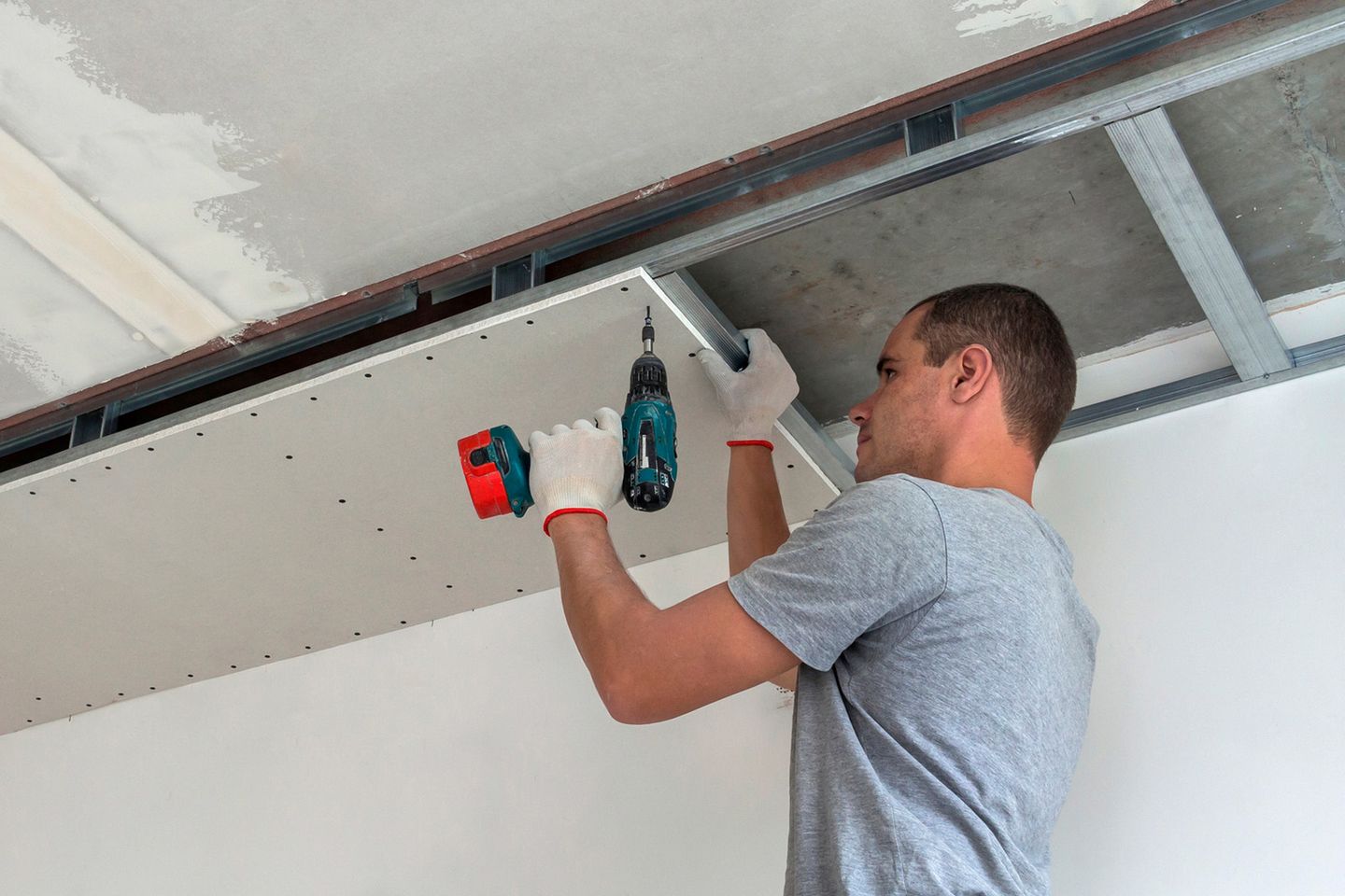 Ein Handwerker befestigt eine Gipskartonplatte an einer Decke mit einem Akkuschrauber, er trägt Handschuhe und ein graues T-Shirt.