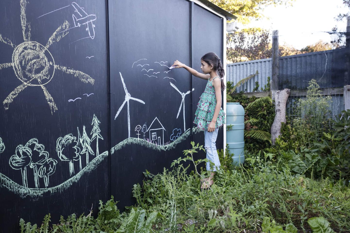 Ein Mädchen zeichnet mit Kreide auf eine große schwarze Wand im Garten. Das Bild zeigt Sonne, Windräder, Bäume, ein Haus und ein Auto – Symbole für Nachhaltigkeit und erneuerbare Energien.
