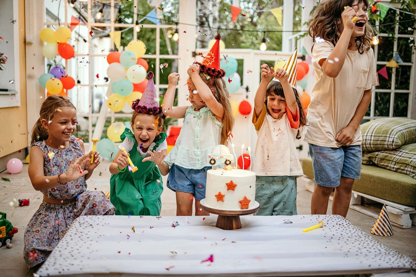 Eine Gruppe Kinder feiert ausgelassen eine Geburtstagsparty mit Partyhüten, Konfetti und einer Torte mit Kerzen.